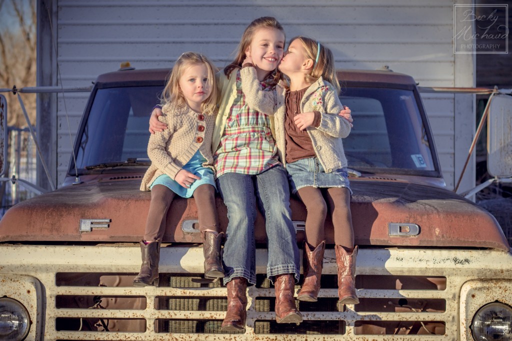 Sisters sitting a on a truck, one girl kisses the oldest girl's cheek