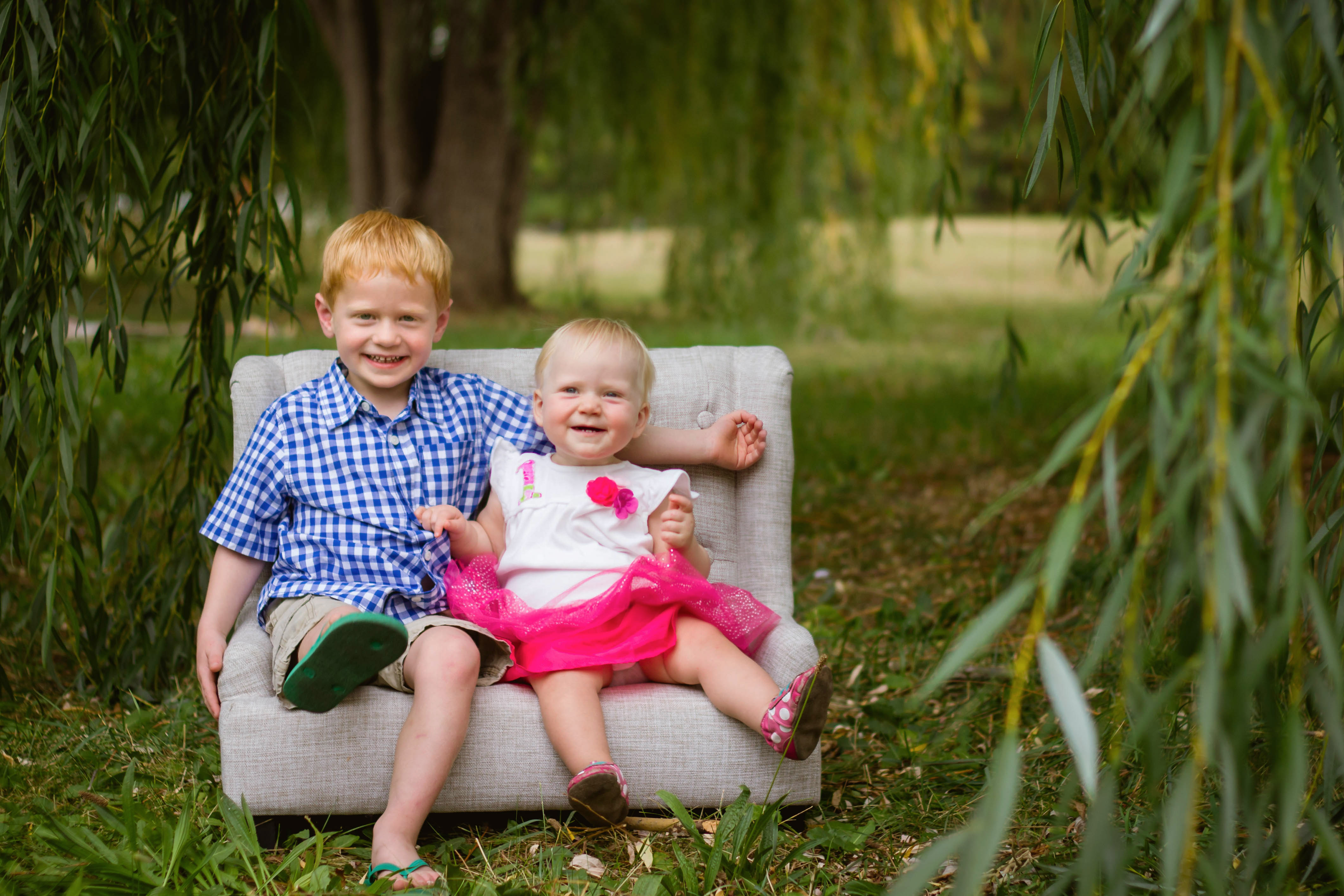 Two siblings play together on a small couch under a willow tree in Fort Collins