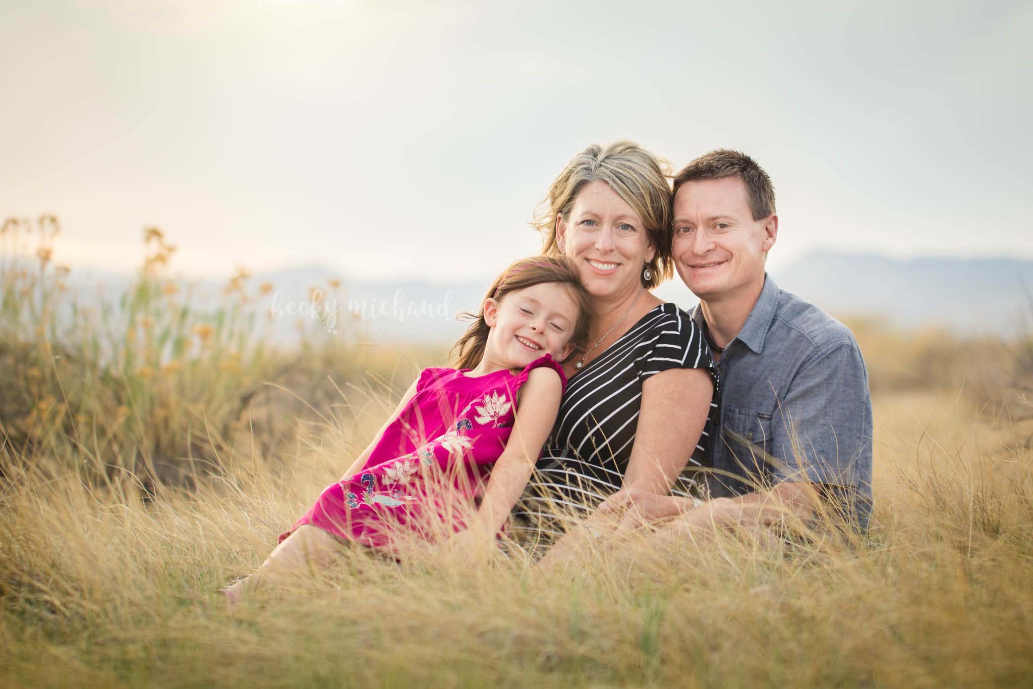 A family of three poses in a field for their photo session with mountains in the background