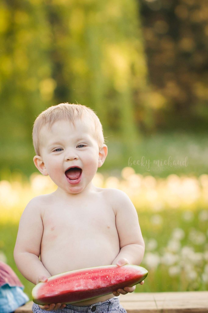 baby boy smiling while holding a watermelon slice in a field in Fort Collins, CO