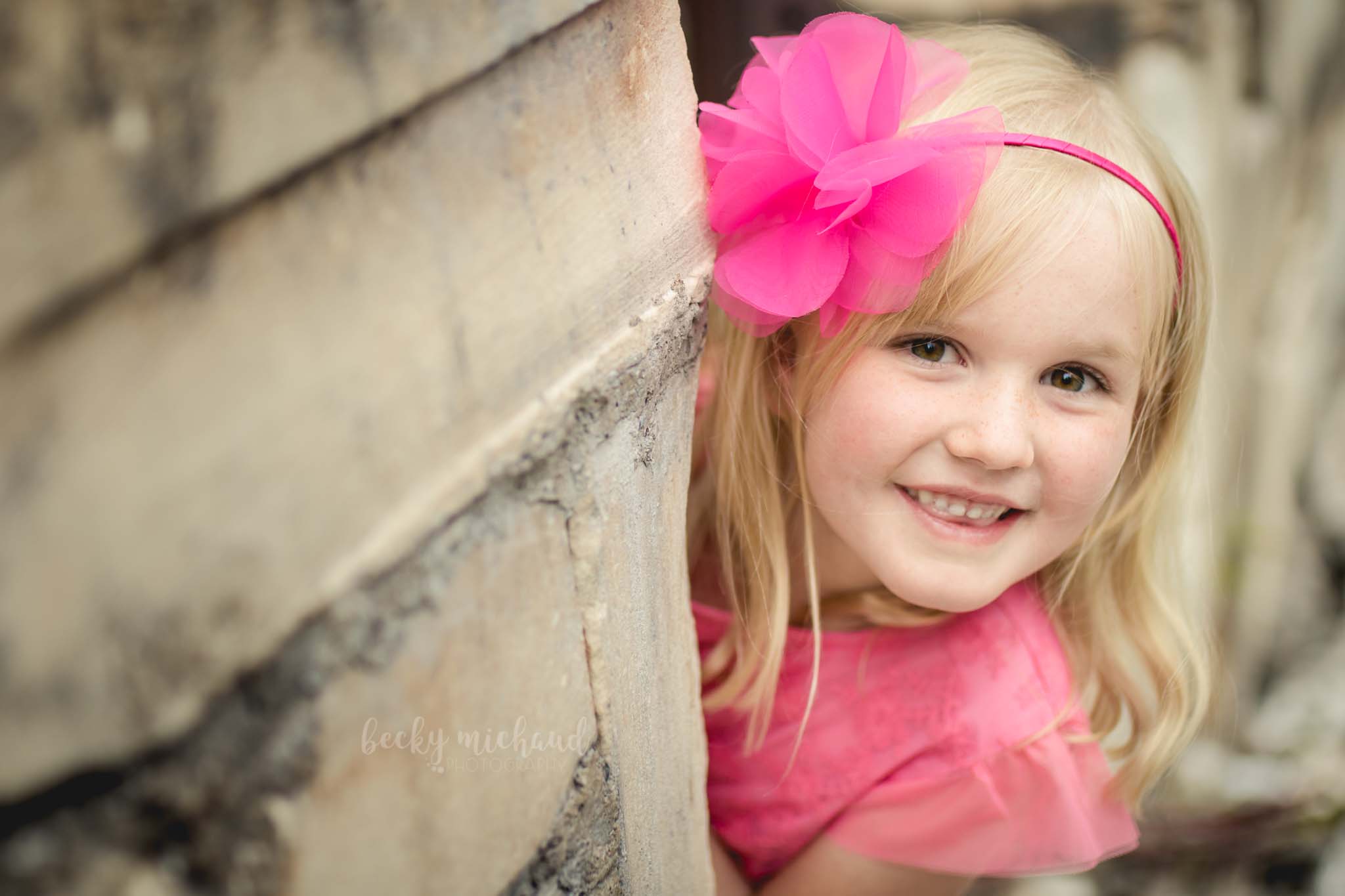 A little girl wearing a pink dress peeks out around a wall