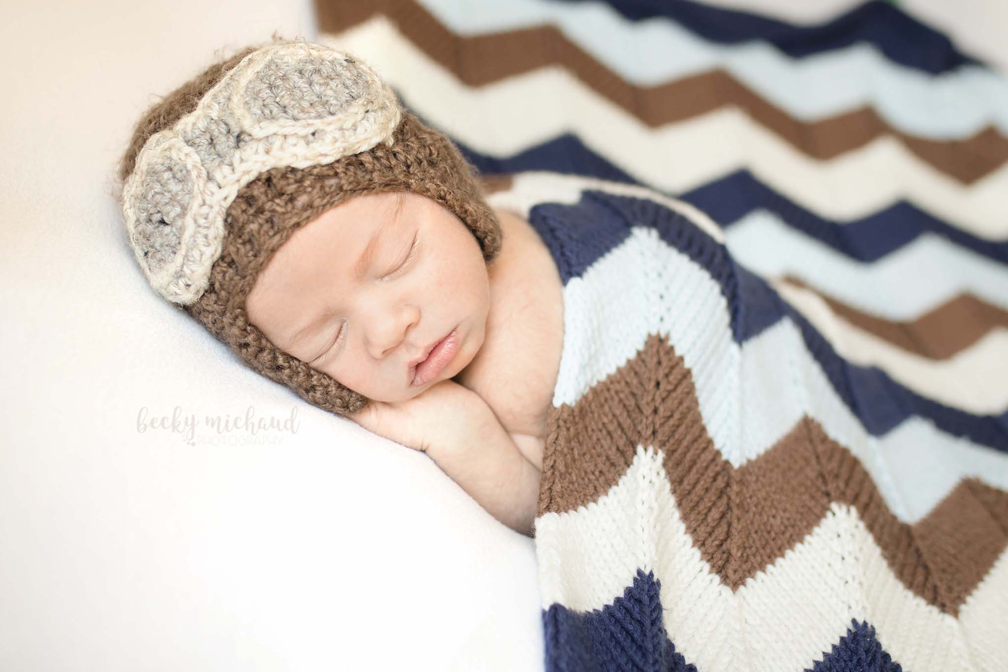 newborn boy cuddled under a chevron knit blanket and wearing an aviator style hat