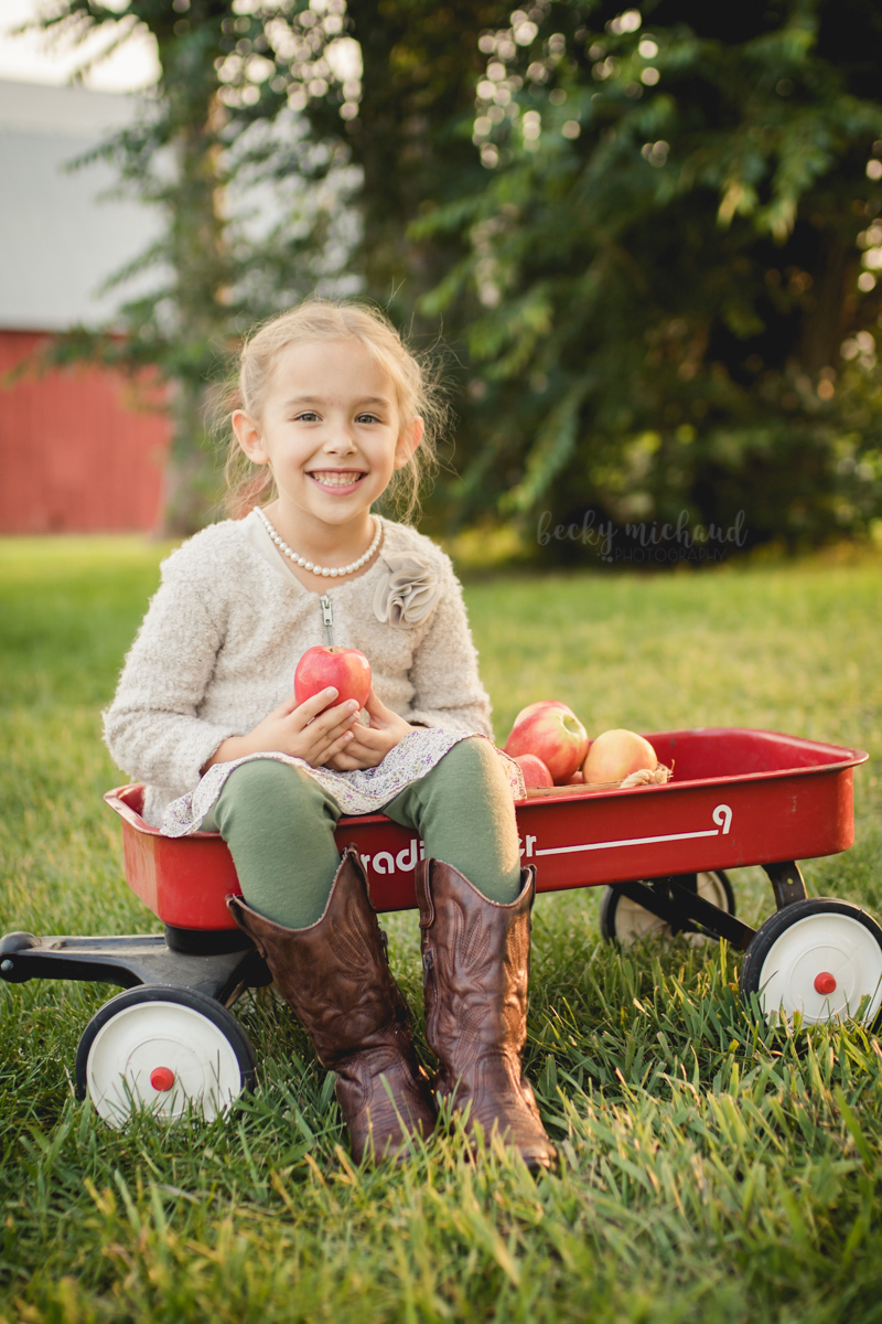 A girl holds and apple in a wagon by a red barn