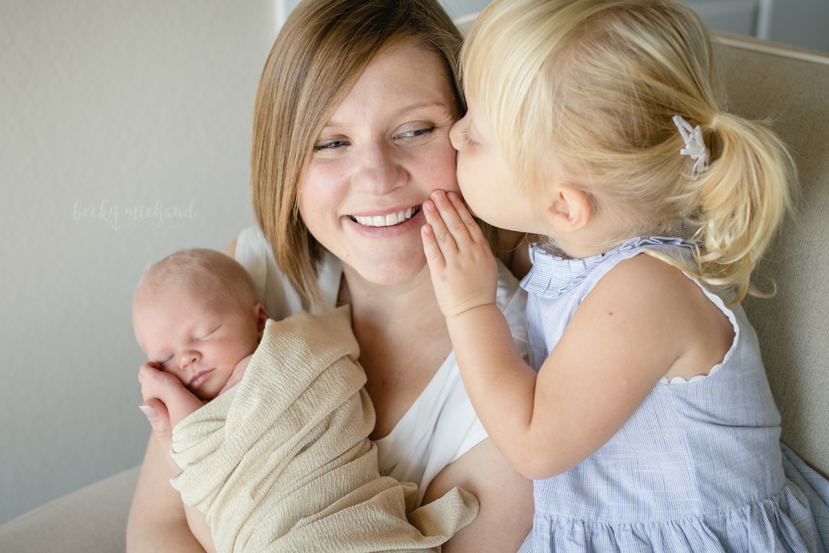 A little girl whispers in her mom's ear while mom holds her baby sister for their in home newborn lifestyle photo session in Fort Collins