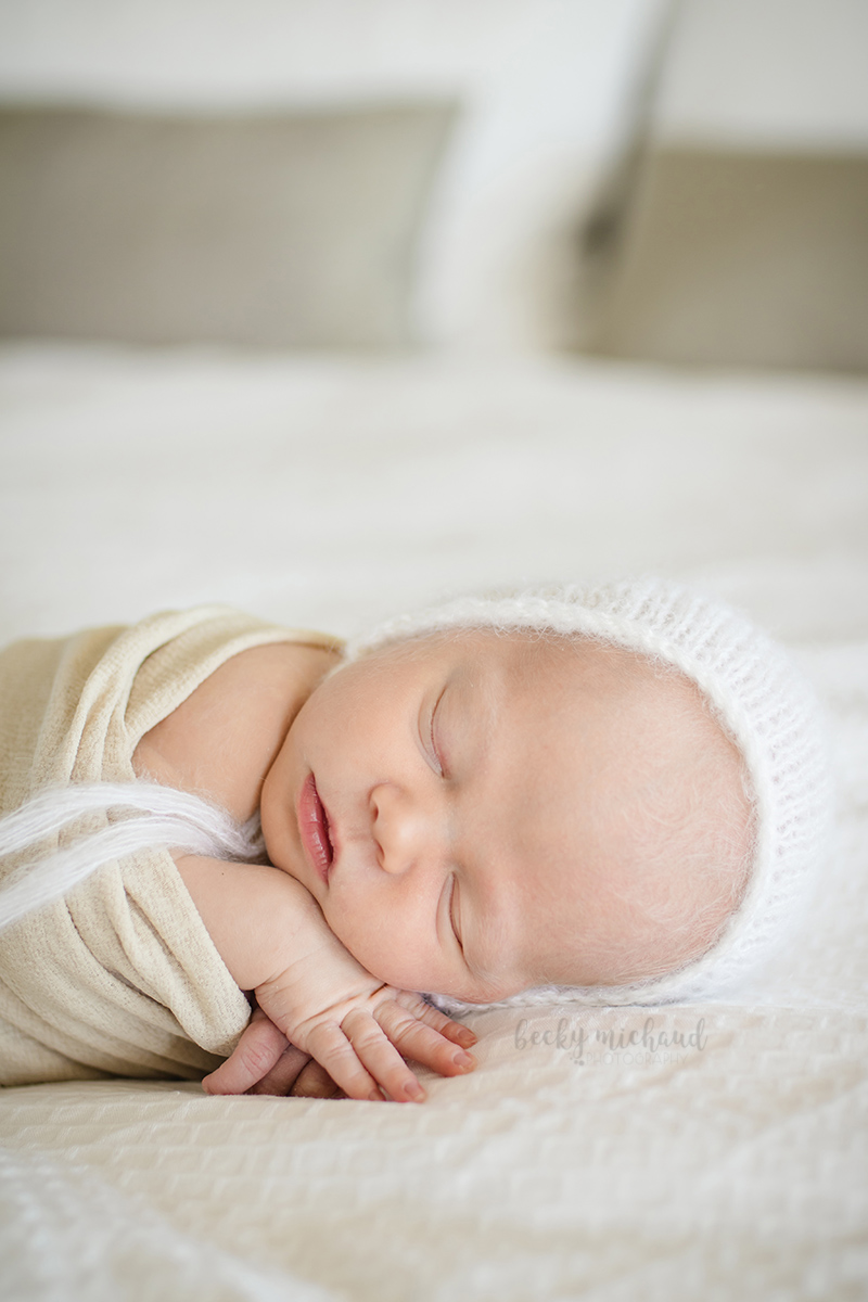 Baby with a white bonnet sleeping peacefully on a bed in her home for her newborn photo session in Fort Collins, CO 