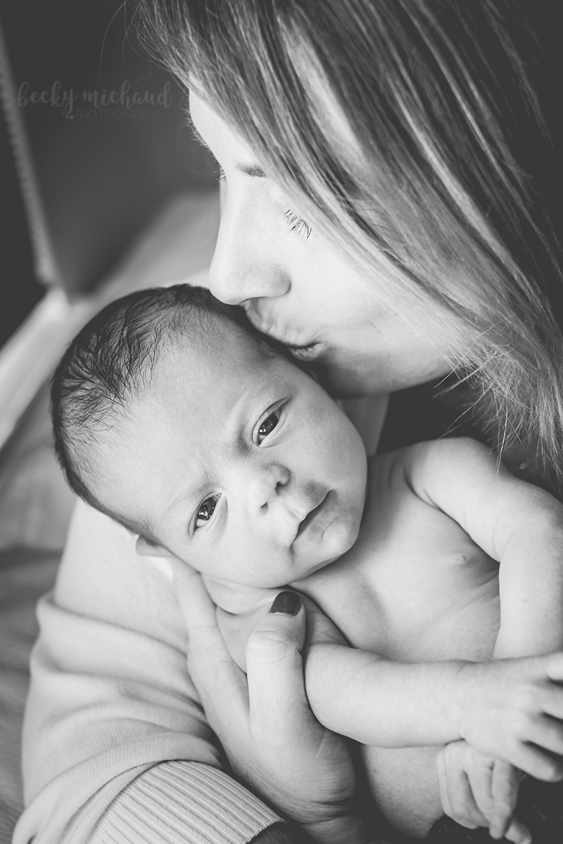 black and white photo of newborn boy making eye contact with the camera while his mom gives him a kiss