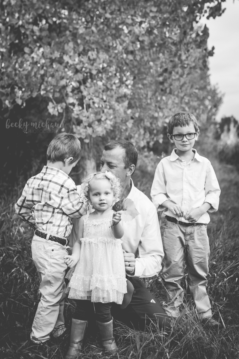 Black and white photo of a little girl holding a leaf while her dad and brothers play in the background in Loveland, Colorado