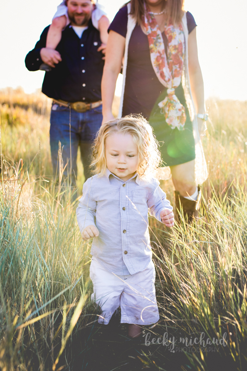 A toddler walks through tall grass with his family behind him