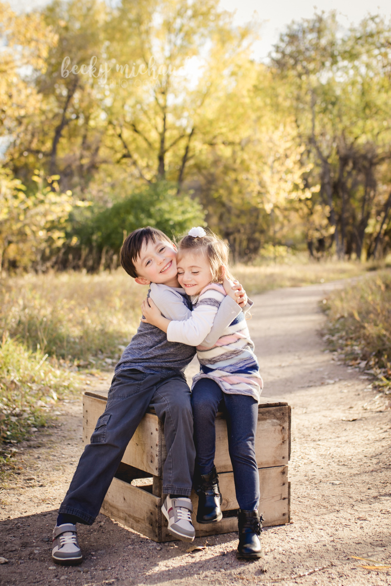 Brother and sister hug while sitting on a crate on a wooded path