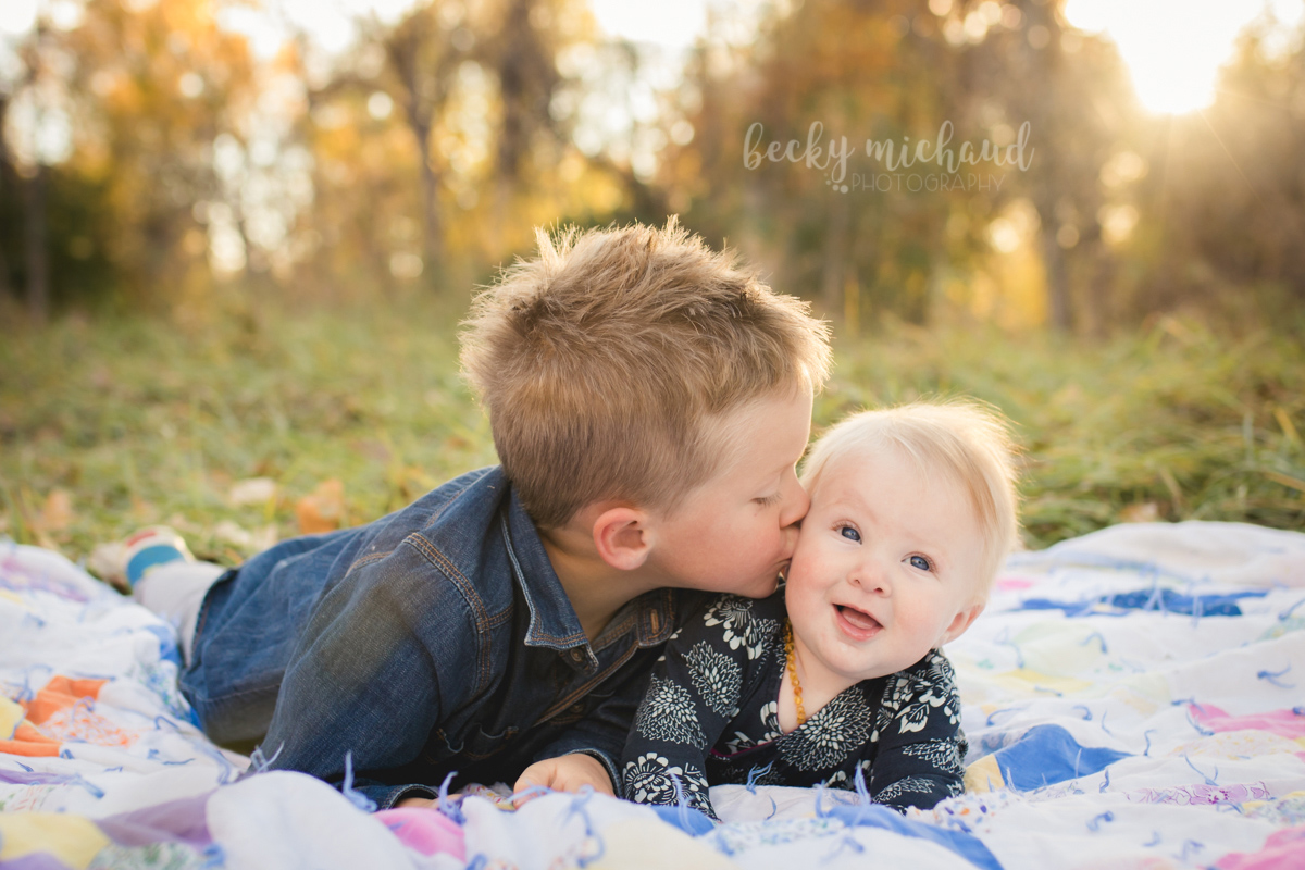 A big brother kisses his baby sister on the cheek while lying on an antique quilt