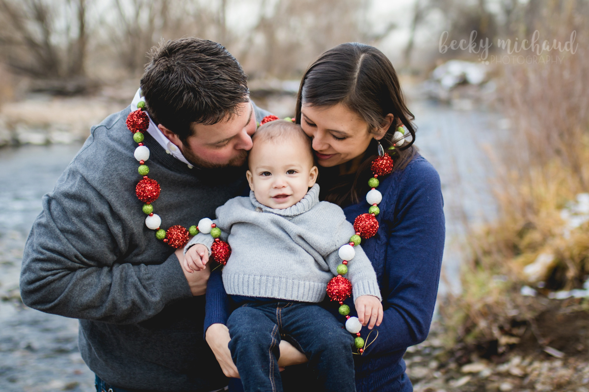 Mom and dad kiss their baby boy while wearing a Christmas garland