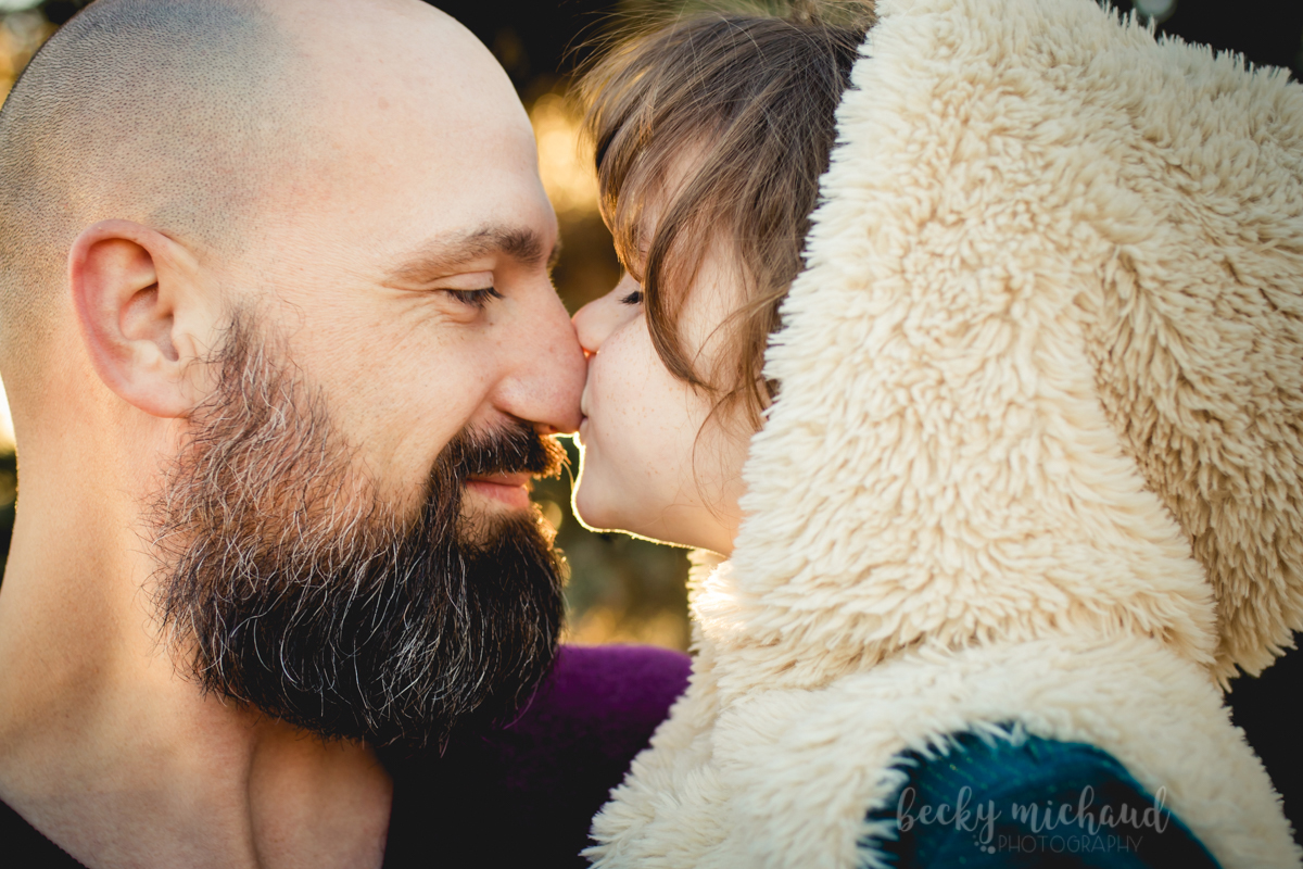 A little girl wearing a furry hood kisses her dad on the nose during their family photo session in Fort Collins