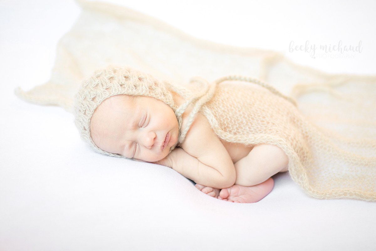 A baby girl poses on a white blanket with a tan bonnet and wrap during her in home Fort Collins newborn session