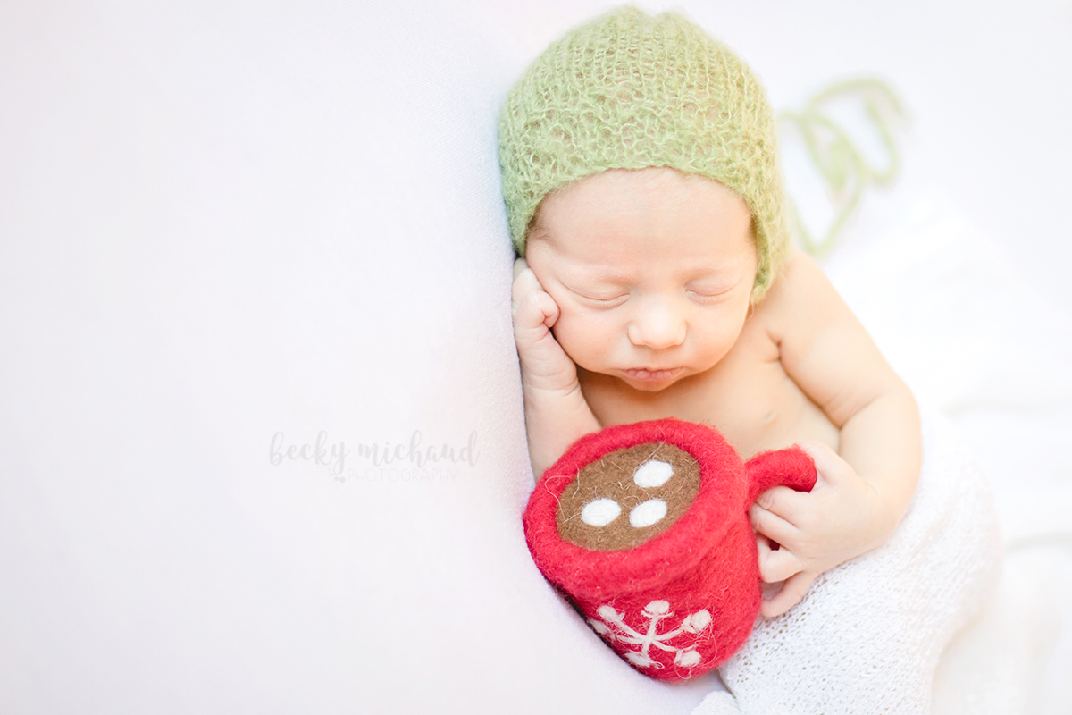 Newborn baby girl holding a felted wool cup of hot cocoa and wearing a green bonnet