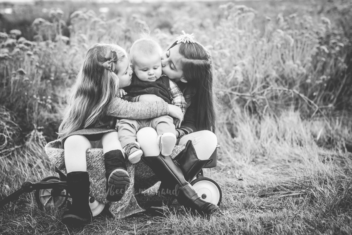 Black and white portrait of two sisters kissing their baby brother on the cheeks