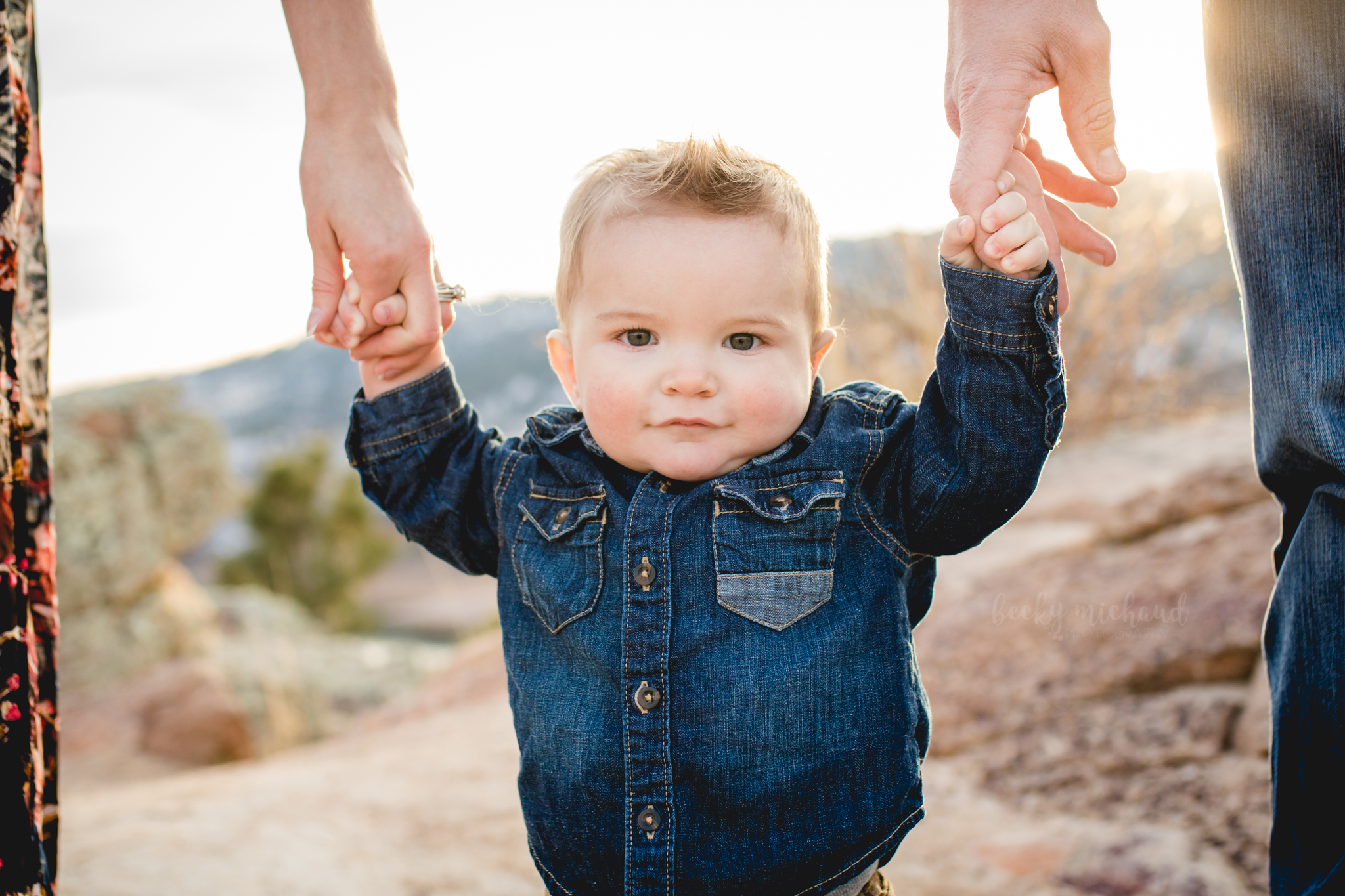 A baby boy holds on to both parents' fingers with the Colorado foothills in the background