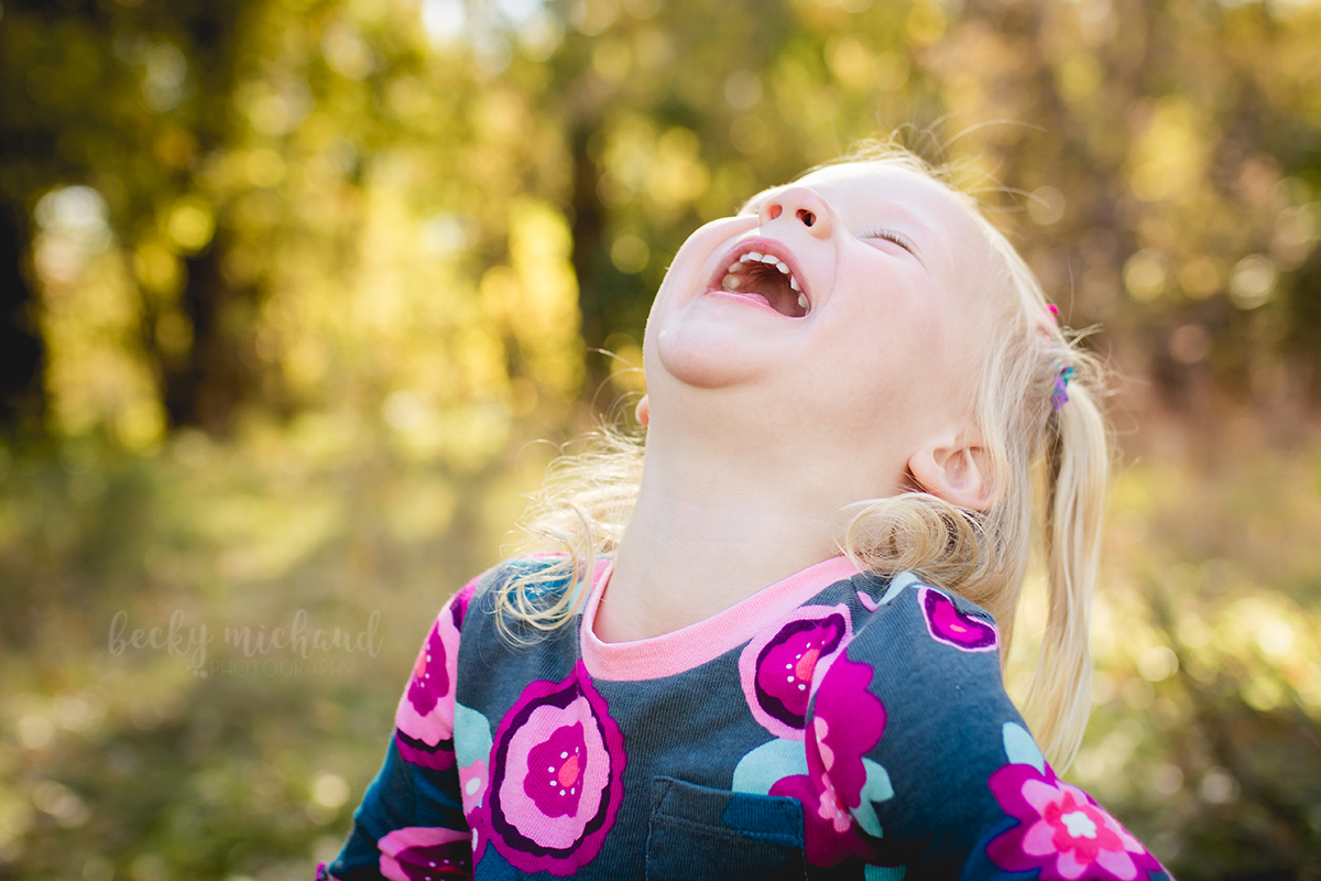A little girl in a pink flowered dress tips her head back and laughs during her Fort Collins family photo session