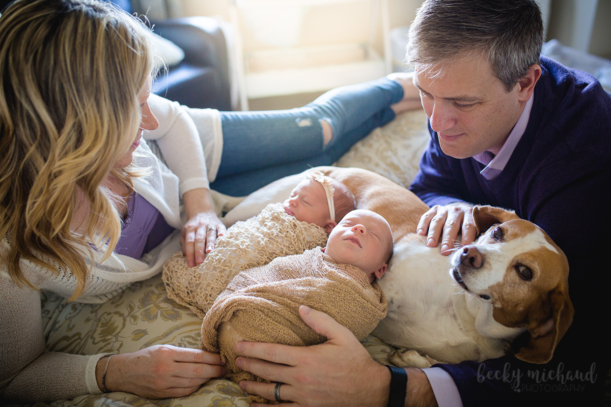A family of two parents, twin newborns, and a beagle lie on a bed together during their in home newborn photo session
