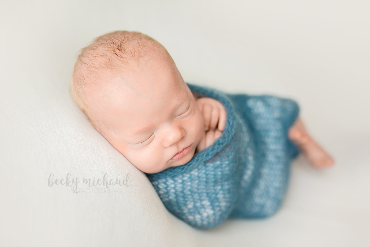 Baby boy wrapped in a blue knit blanket lying on a white backdrop for his Fort Collins newborn photo session