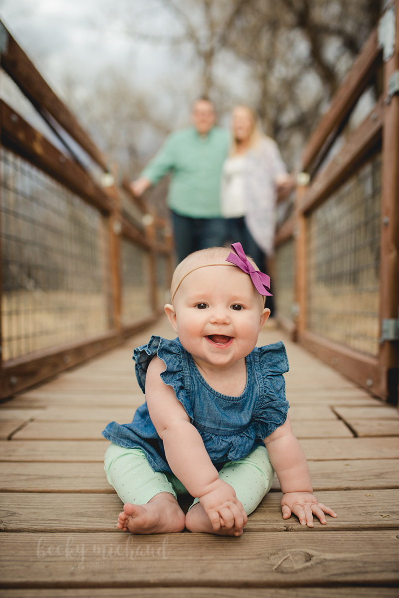Fort Collins photography of a baby girl sitting on a bridge with her parents behind her