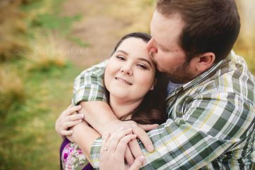 A woman smiles at the camera while her fiance kisses her on her head during her photography session in Longmont Colorado