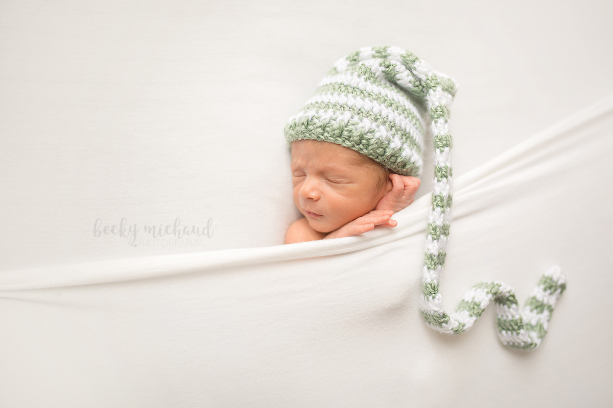 A baby poses for his newborn photos with a green and white hat tucked under a white blanket