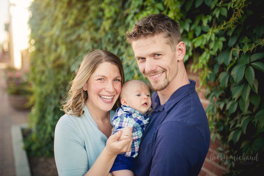 A family of three smiles together in front of a wall of ivy in Old Town, Fort Collins