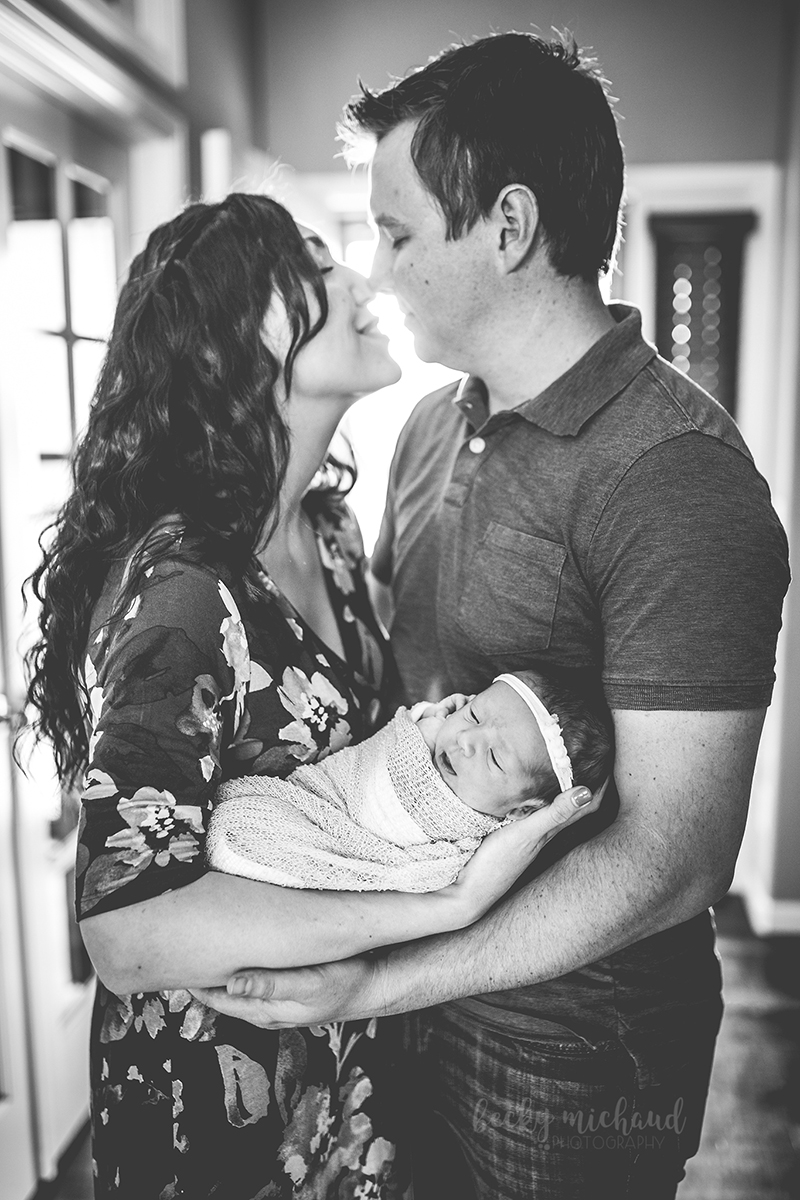 black and white photo of new parents embracing as they hold their baby girl in their backlit entryway