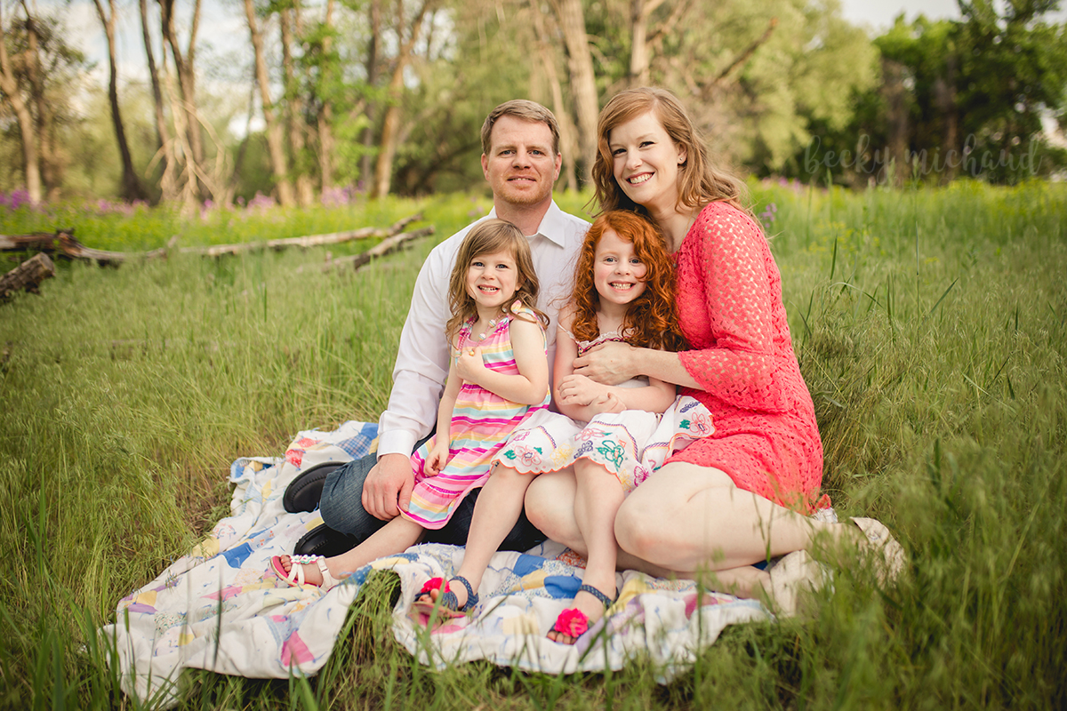 A family of four sits on a blanket in a field during their family portrait session in Fort Collins