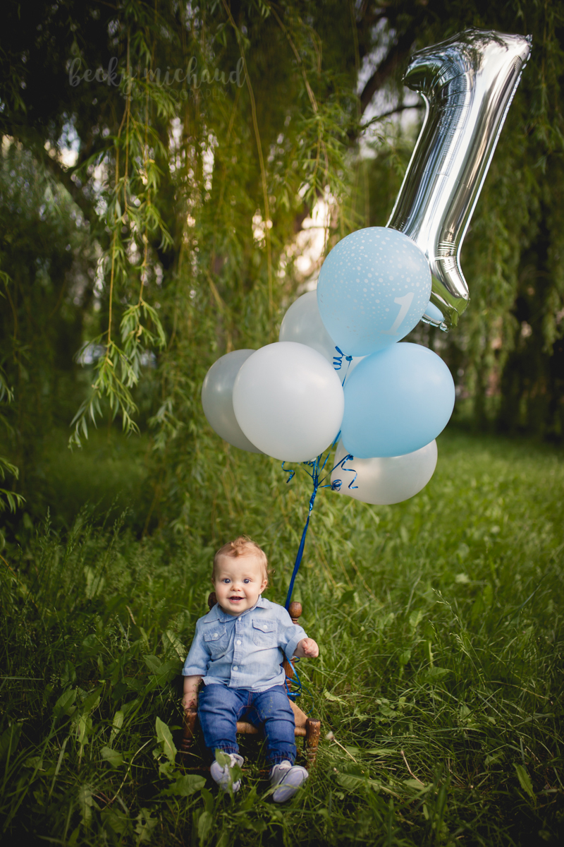 One year old photo of a little boy sitting in a wooden chair with a big bouquet of balloons
