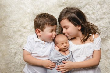A brother and sister kiss their baby brother during their newborn photo session in their Fort Collins home