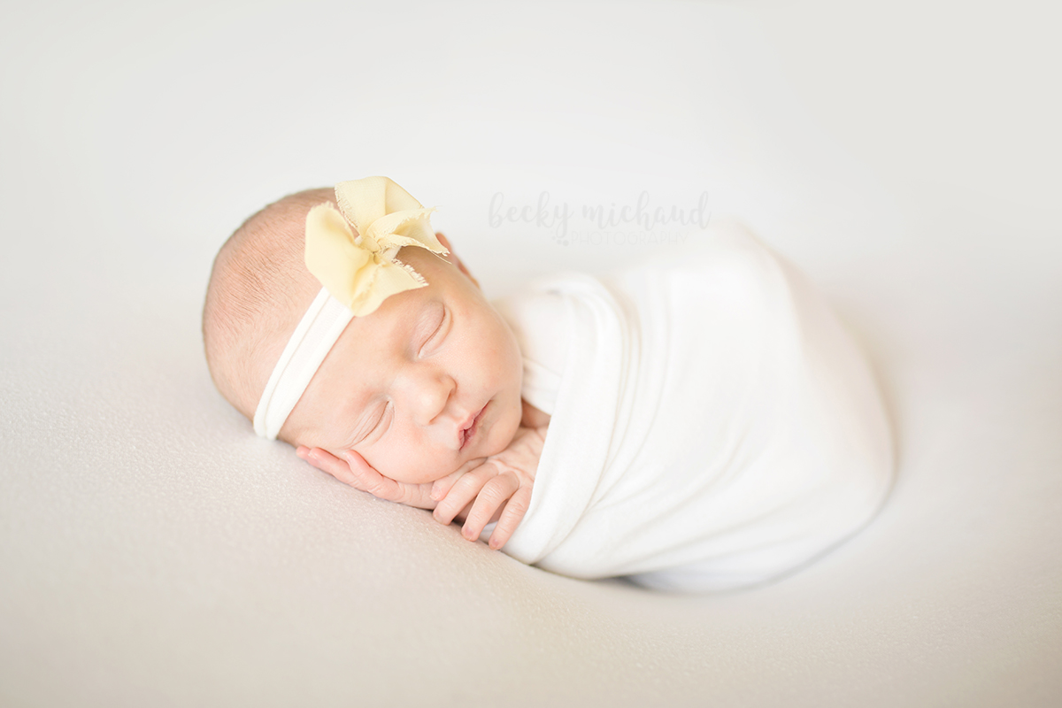 Simple newborn photo of a baby girl in a white wrap with a yellow bow, taken in her Timnath, Colorado home