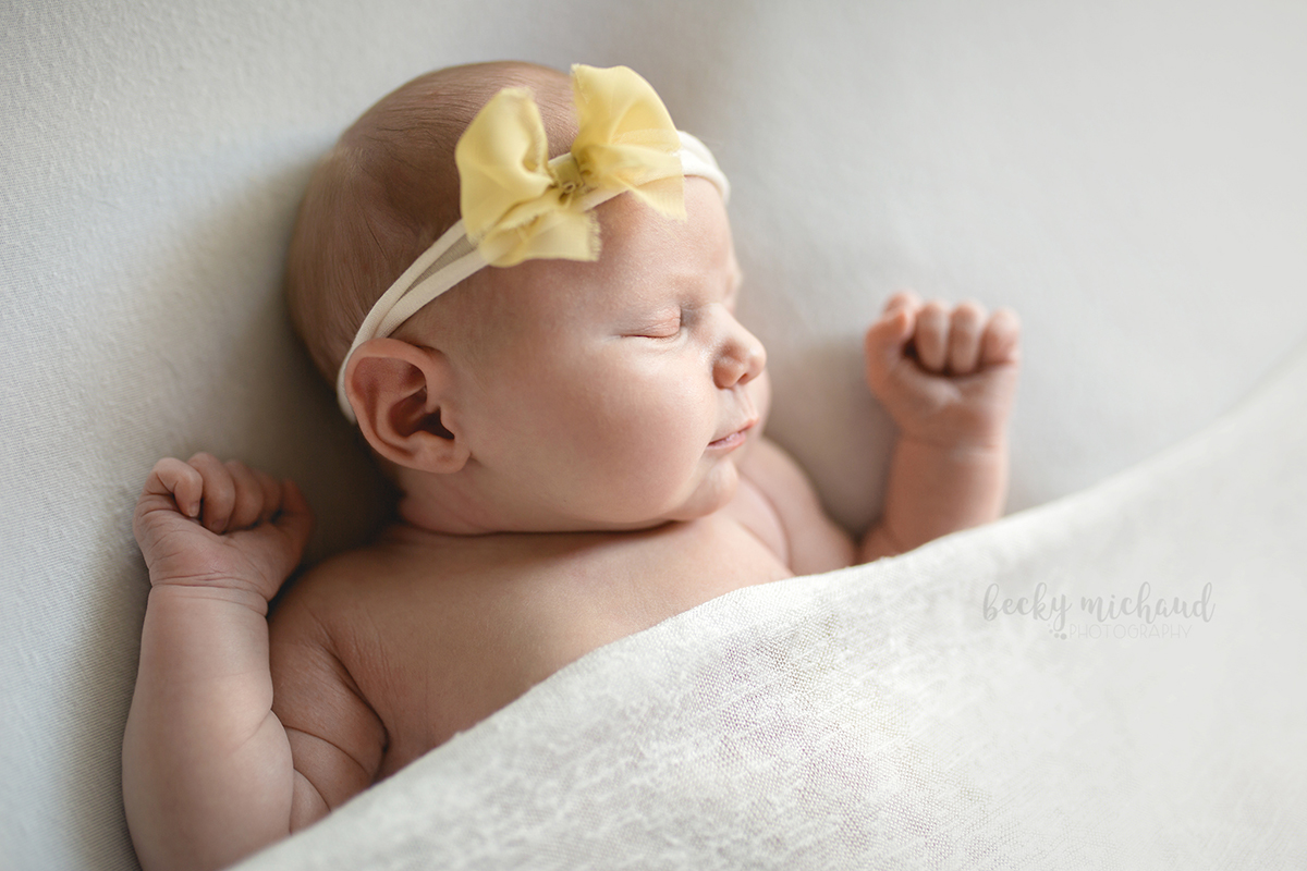A baby girl is tucked in under a plain white blanket with a yellow bow in her hair for her newborn photo session in her Fort Collins home