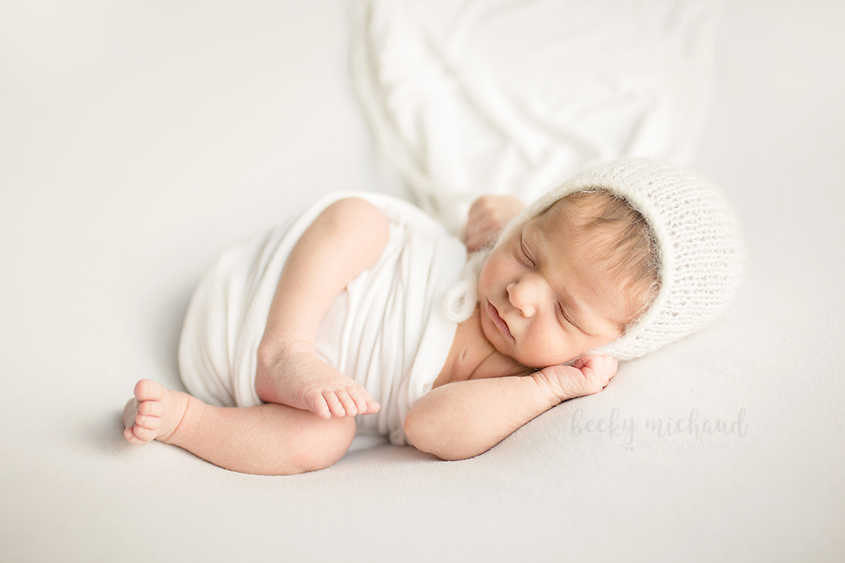 Curled up newborn baby in a white wrap and bonnet in a posed photo in her Timnath, Colorado home