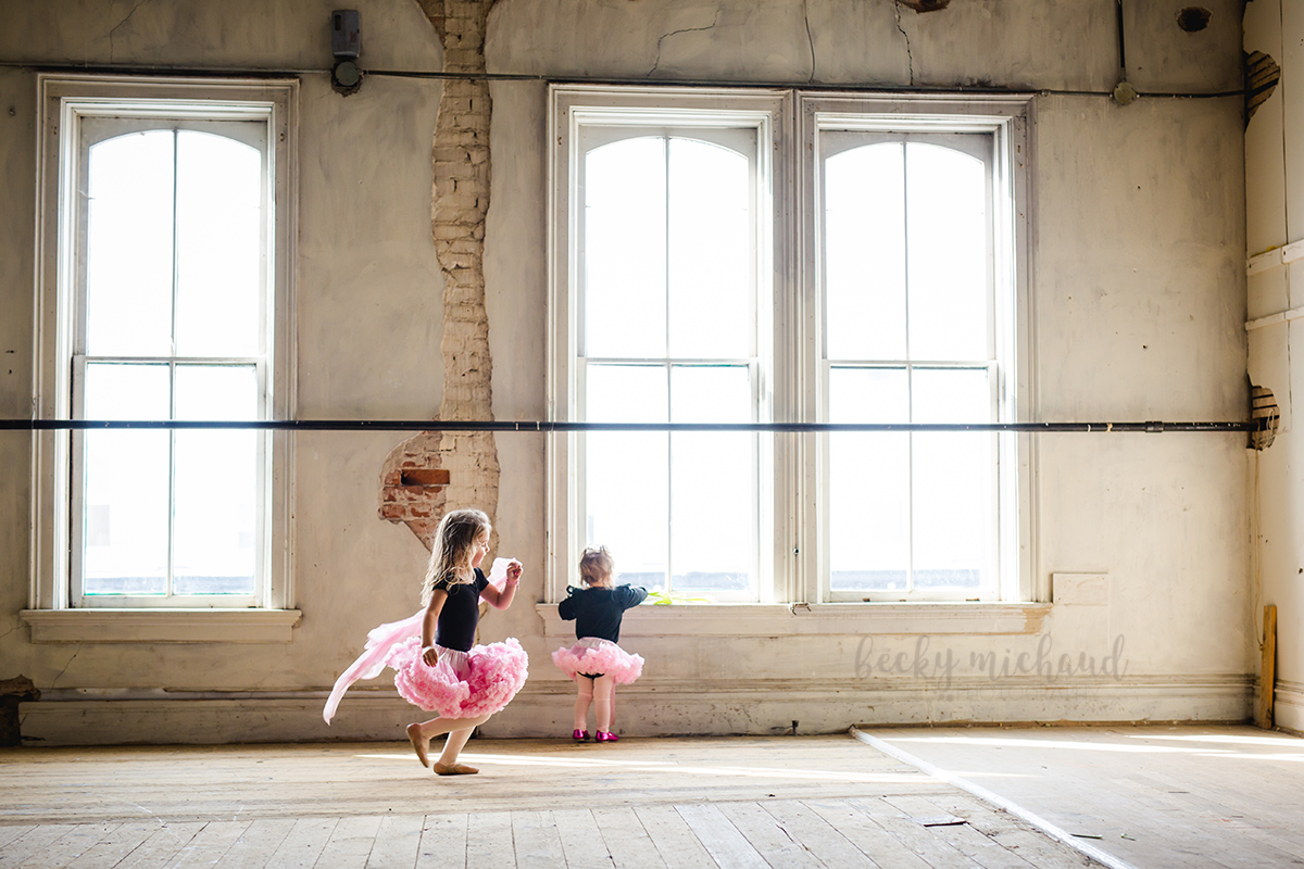 Two little dancers wearing tutus run around a big empty room in an Old Town Fort Collins building