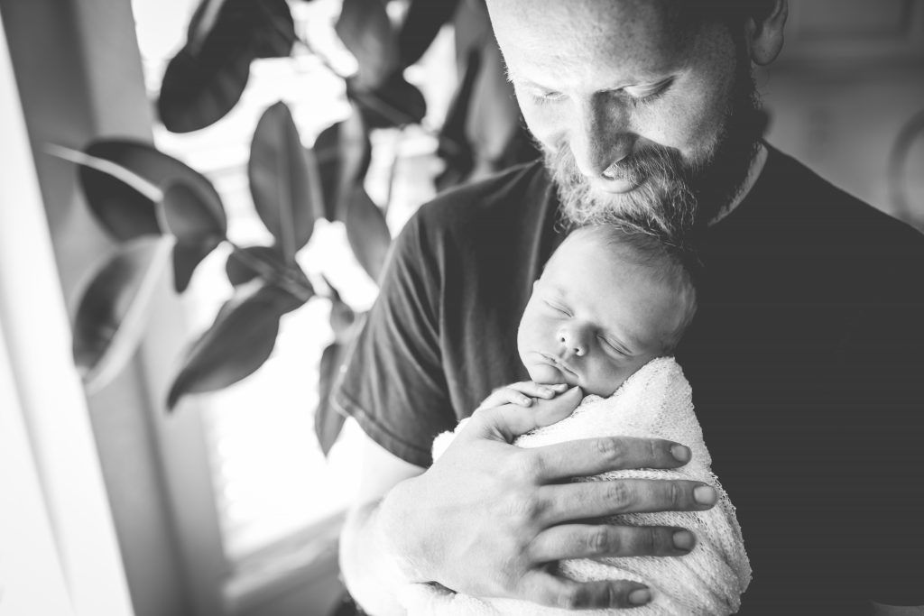 A proud dad looks down on his newborn boy in a black and white photo taken near a window in their Fort Collins home
