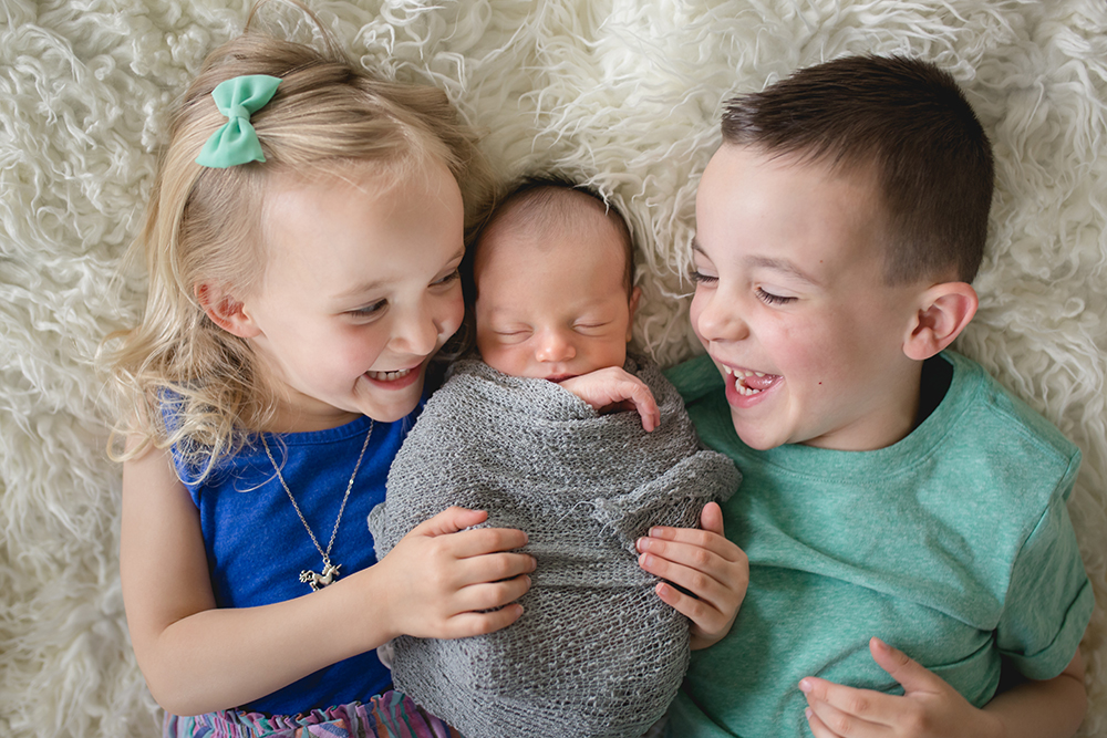 A brother and sister laugh together while snuggling their newborn brother during their in home newborn photo shoot