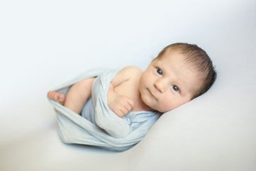 A wide awake newborn boy looks at the camera as he poses on a white blanket in his Johnstown, Colorado home
