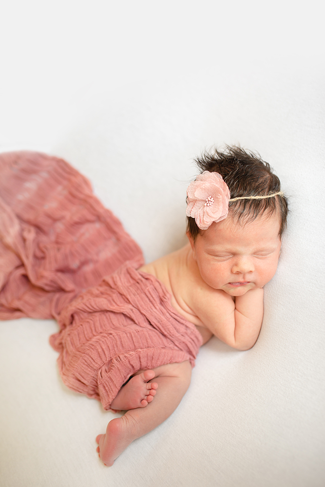 Photo of a baby girl laying on a white blanket with a pink wrap and pink flower headband