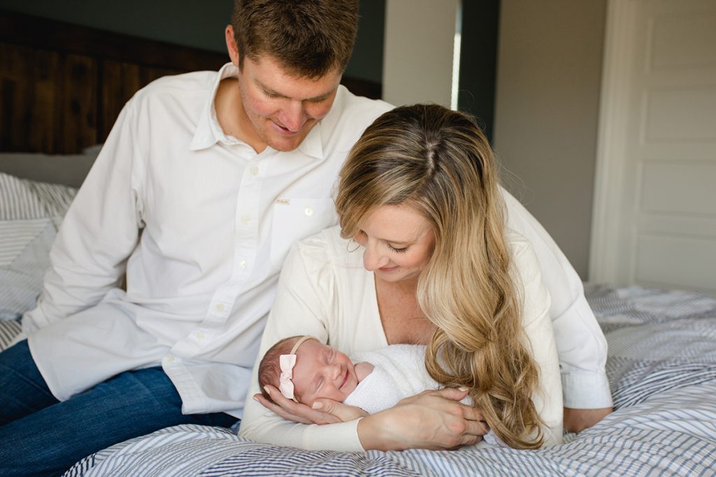 A newborn baby girl smiles as her parents hold her on their bed in a lifestyle photo taken in Loveland, Colorado