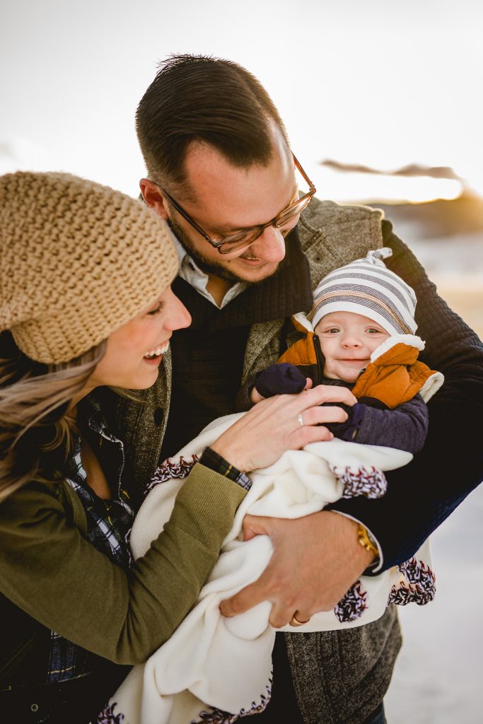 portrait of a baby smiling at the camera while his mom and dad snuggle with him in the Colorado snow