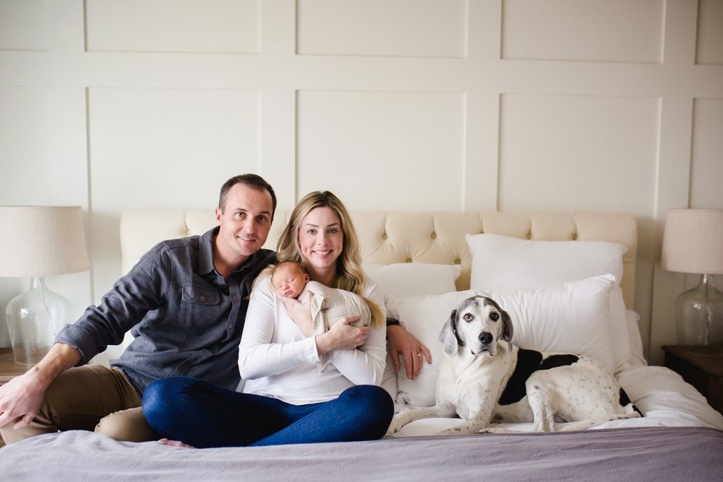 New parents pose with their newborn and their dog on their bed during their newborn photo session in their Colorado home
