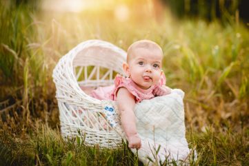 Baby girl plays with her tongue as she sits in a white wicker basket in a photo taken by Becky Michaud, Colorado baby photographer