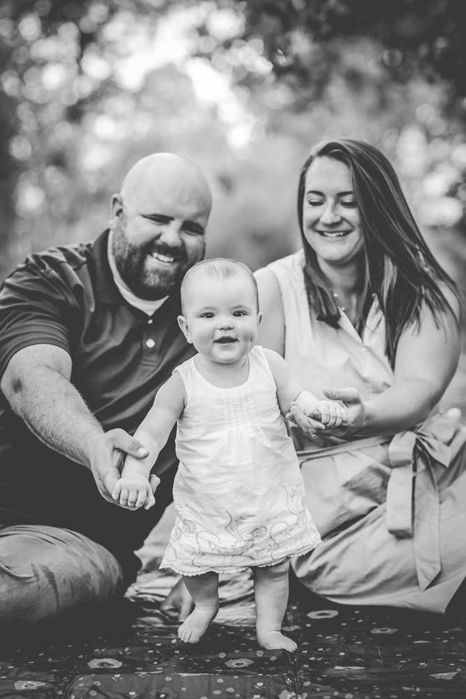Black and white photo of a mom and dad holding the hands of their baby girl as she smiles for the camera