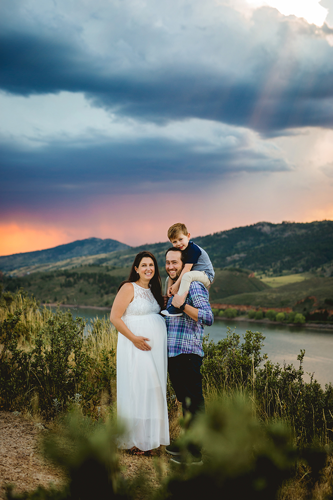 A family poses for their maternity photos at Horsetooth Reservoir with a bright pink sunset behind them