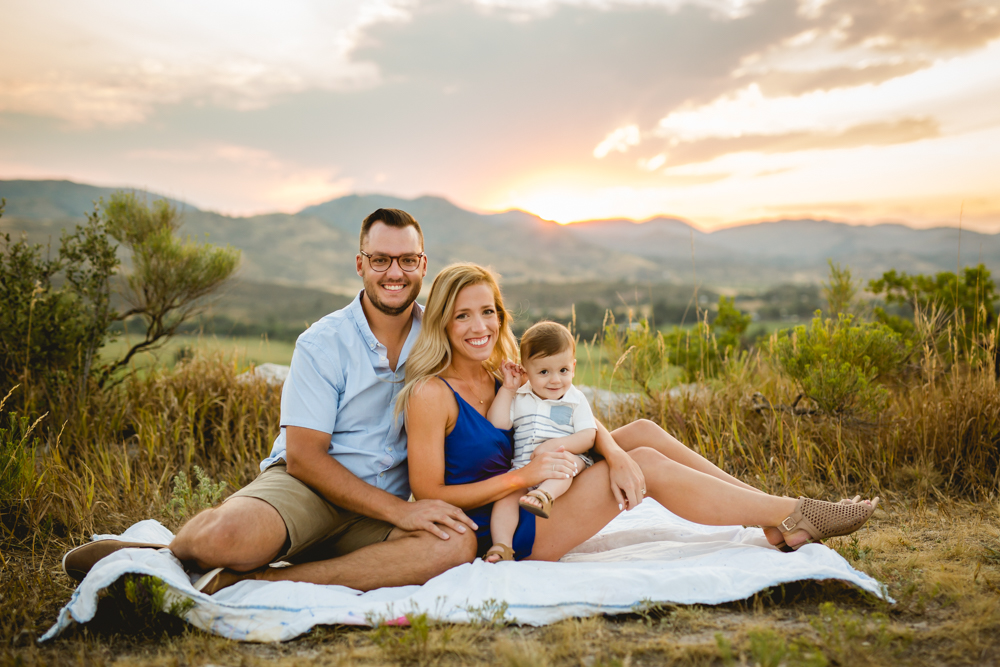 A family of three sits on a hilltop overlooking Bellvue, Colorado as the sun sets behind them