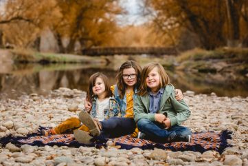 Three siblings sit by the river in fall in a photo taken by Becky Michaud, Fort Collins photographer