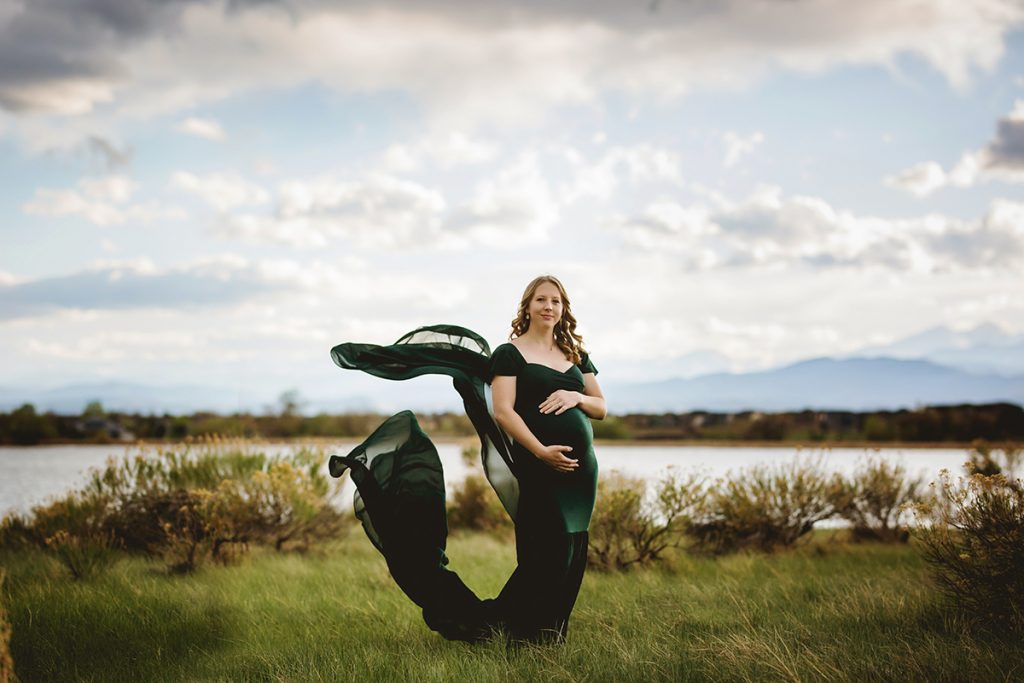 A mom in the Chicaboo Monroe gown poses by a lake with the Colorado mountains in the background