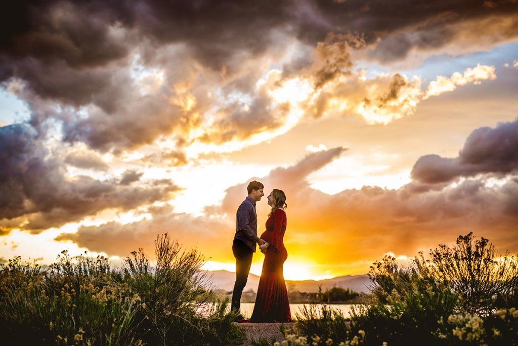 A couple stands by a lake in Loveland, Colorado as the sun sets behind them during their maternity photo shoot with Fort Collins photographer Becky Michaud