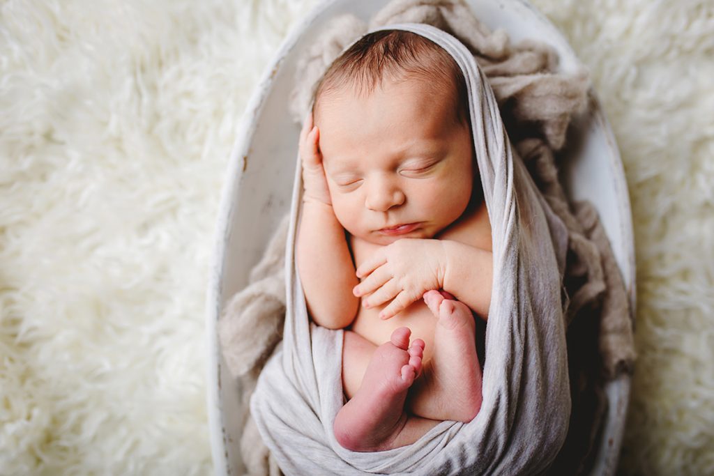 Newborn baby curled up in a wooden bowl for his newborn photos session in his Wellington, Colorado home